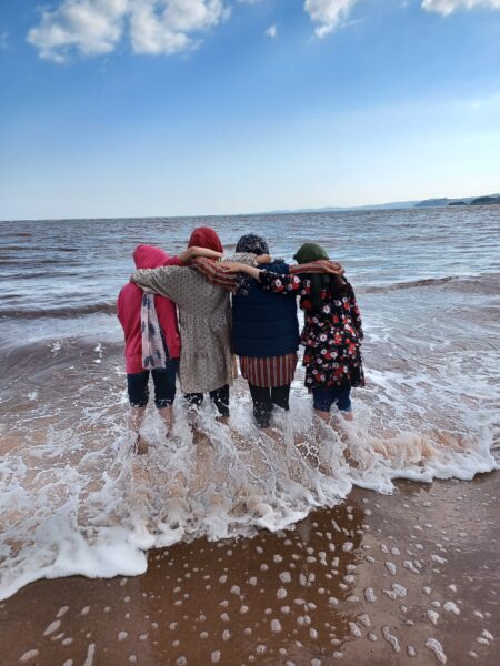 A group of four afghan women at the beach standing in the water linking arms and hugging as they look out at the ocean.