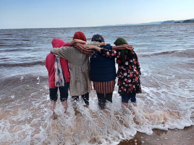 A group of four afghan women at the beach standing in the water linking arms and hugging as they look out at the ocean.