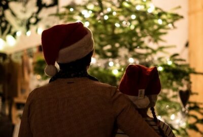 Older person and a young person stood looking at a Christmas tree. Both are wearing Father Christmas red hats.