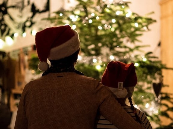 Older person and a young person stood looking at a Christmas tree. Both are wearing Father Christmas red hats.