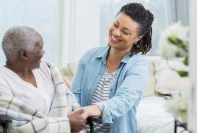 A female care worker, smiling, holding the hand of the person they are caring for.