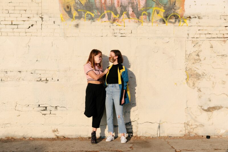 Two young people stood side by side with their back to a wall, talking to each other.