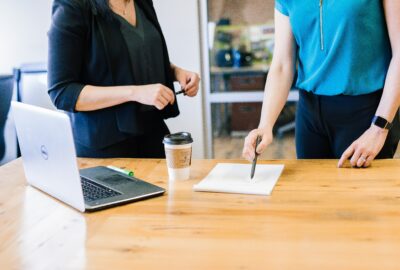 two people stood working at a desk, with a laptop in front of them on the desk.