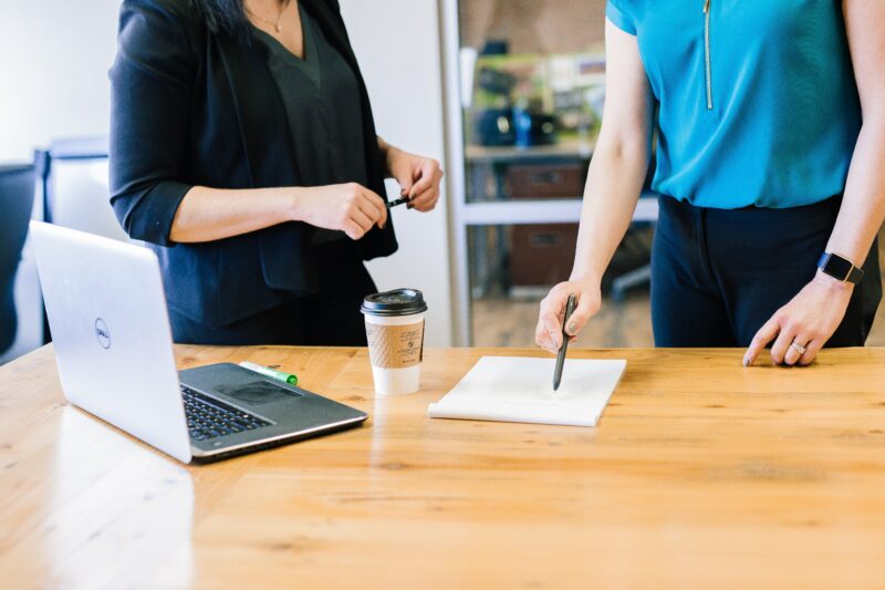 two people stood working at a desk, with a laptop in front of them on the desk.