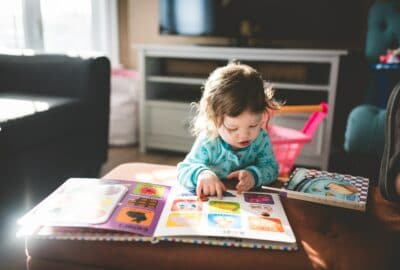 A young girl stood at a low table with a young child's reading book on the table. She is looking at the pictures.