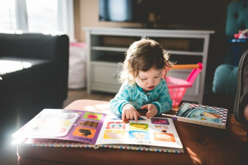 A young girl stood at a low table with a young child's reading book on the table. She is looking at the pictures.