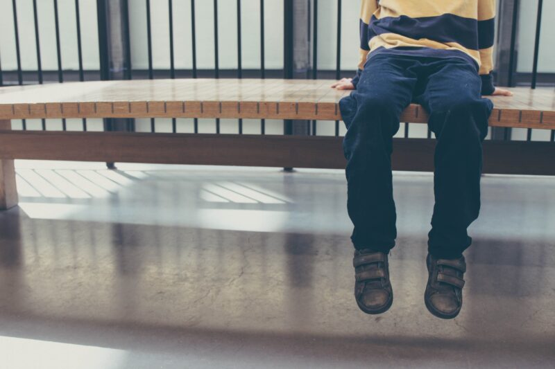 A anonymous child sat on an indoor bench, against some railings