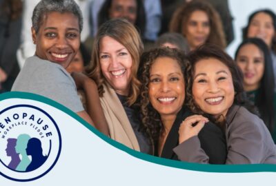 Four women huddling close and smiling at the camera, in front of a larger group of women