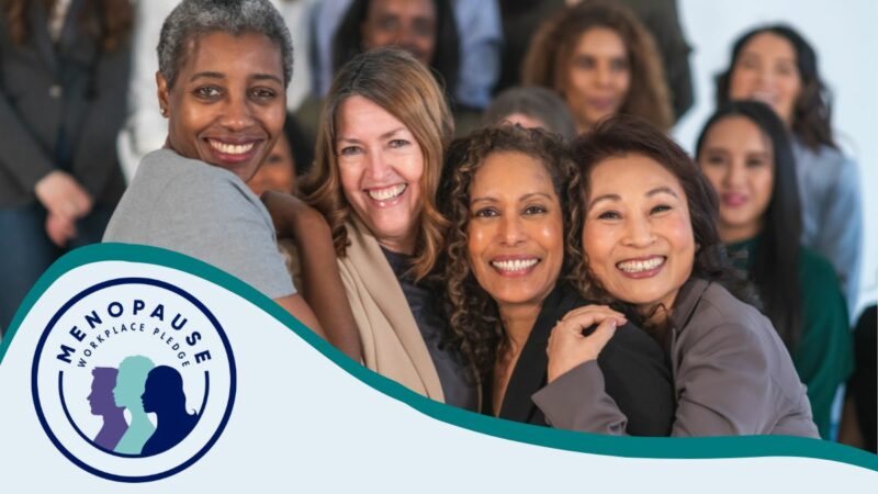 Four women huddling close and smiling at the camera, in front of a larger group of women