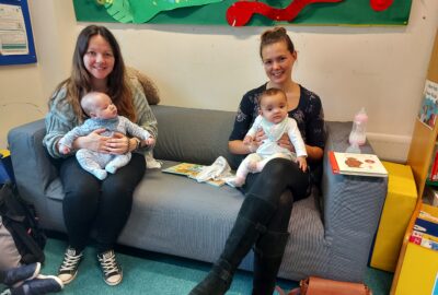 Two mums with young babies on their knees sat on a settee in the children's section of Tiverton Library