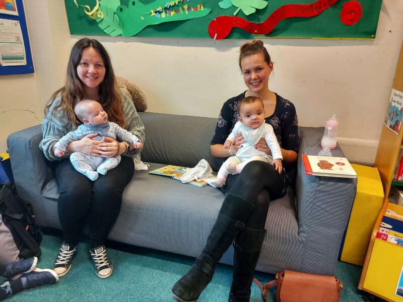 Two mums with young babies on their knees sat on a settee in the children's section of Tiverton Library