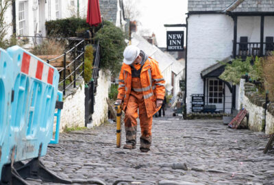 A broadband engineer wearing orange overalls and hard hat, walking up cobbled street in Clovelly