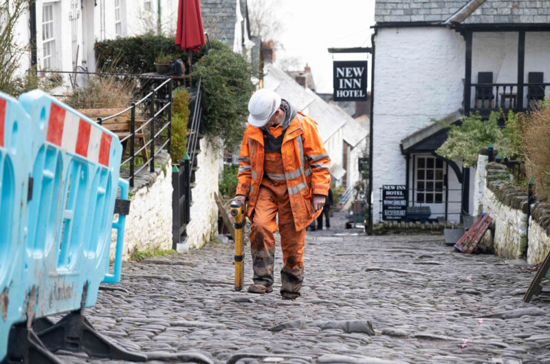 A broadband engineer wearing orange overalls and hard hat, walking up cobbled street in Clovelly