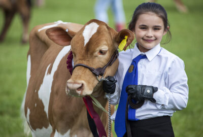 Young girl stood wearing shirt and tie alongside a calf, smiling.
