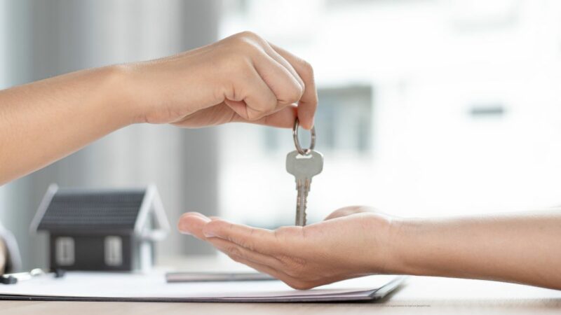 A person handing over some keys to someone else. Only their forearms and hands, and a key dangling from a finger, can be seen.
