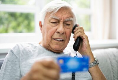 An elderly man talking on the telephone, holding and reading from his bank card.