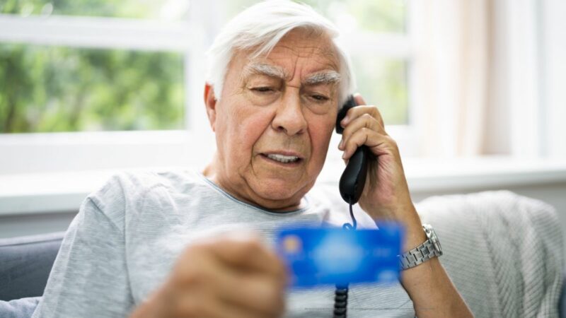 An elderly man talking on the telephone, holding and reading from his bank card.