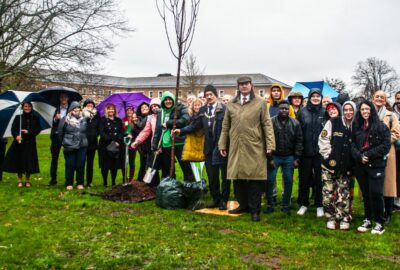 A group comprising young people, councillors and council staff, planting a tree in the grounds at County Hall