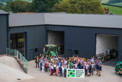 A crowd of people stood looking up at the camera, outside an agricultural building, with a tractor