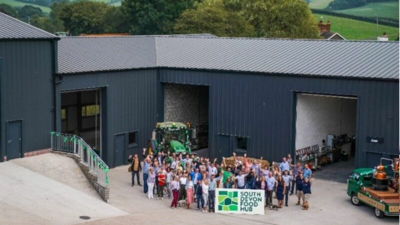 A crowd of people stood looking up at the camera, outside an agricultural building, with a tractor