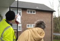 A man in high-vis jacket talking to an elderly lady at her doorstep and pointing up at her roog