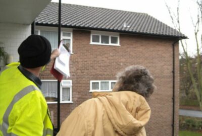 A man in high-vis jacket talking to an elderly lady at her doorstep and pointing up at her roog