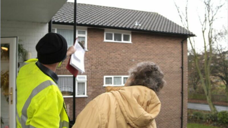 A man in high-vis jacket talking to an elderly lady at her doorstep and pointing up at her roog
