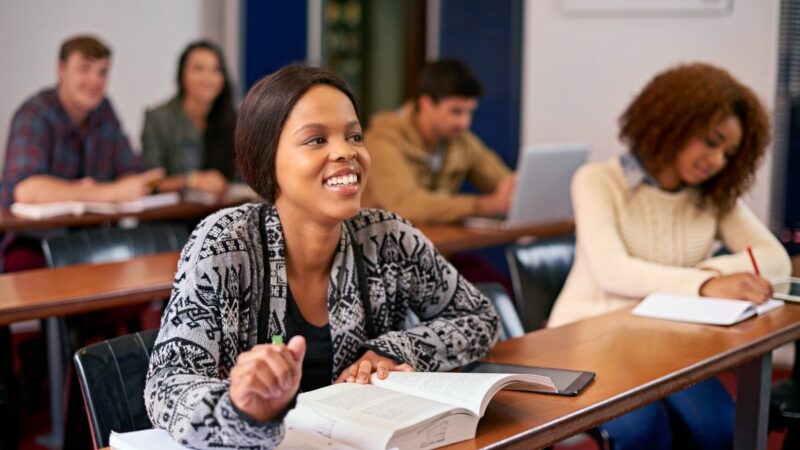 Students in the classroom at college, with books in front of them on their desks, looking up and to the front at their tutor