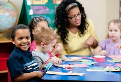 A group of pre-school age children sat around a desk with a pre-school worker engaging with them at a table