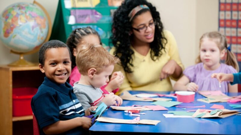 A group of pre-school age children sat around a desk with a pre-school worker engaging with them at a table