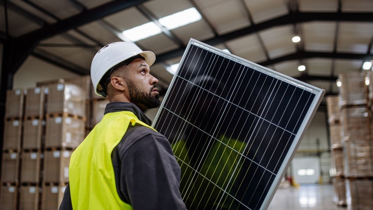 A man in high vis and a hard hat, in a warehouse, carrying a solar panel, with lots of boxes in the warehouse