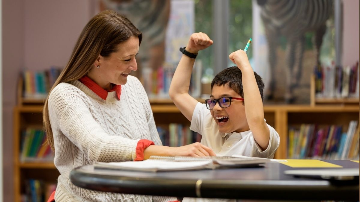 one to one support from an adult with a vulnerable child. The two are sat at a desk with a book in front of them. The child is laughing and smiling.