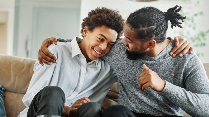 Father with his arm around son sat on sofa