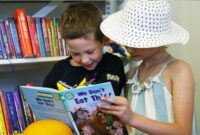 Two young children enjoying looking at a book together in front of book shelves at a library