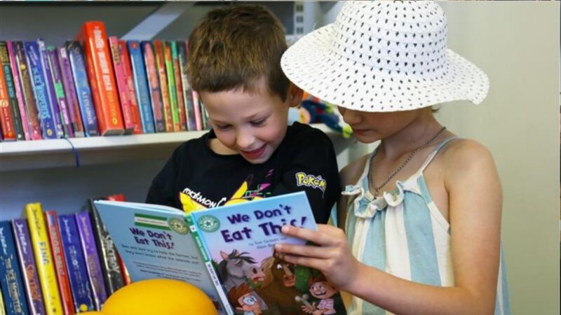 Two young children enjoying looking at a book together in front of book shelves at a library