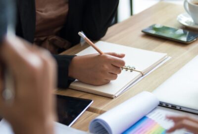 Close up photograph of a meeting desk, with a person writing in their note pad.