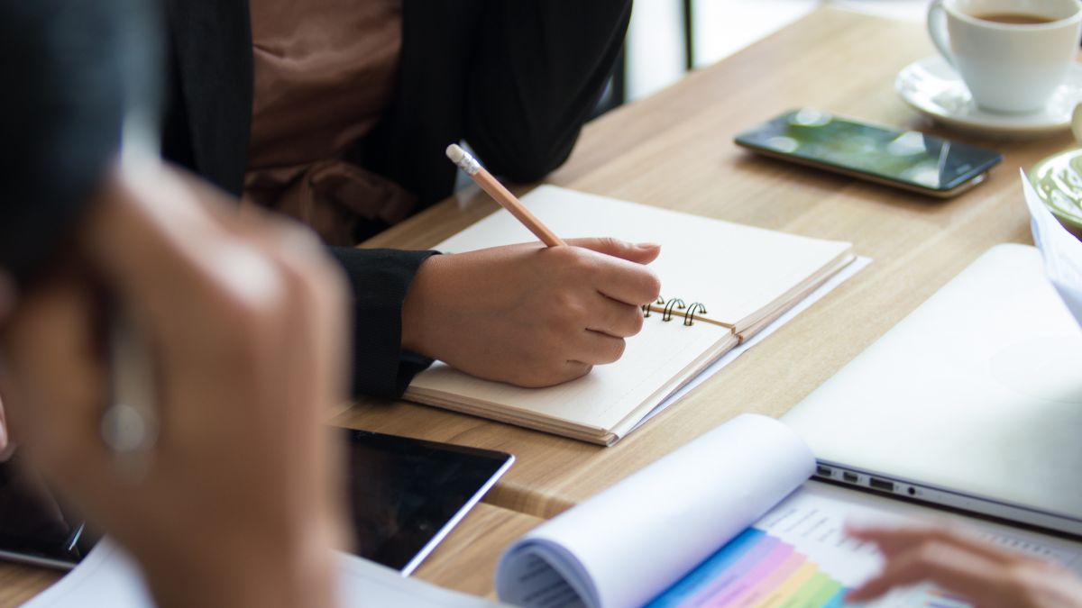 Close up photograph of a meeting desk, with a person writing in their note pad.