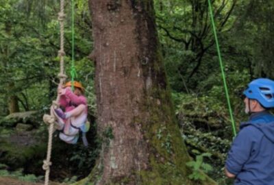 A young girl swinging from a rope swing, supported by a rope and a member of staff holding the rope