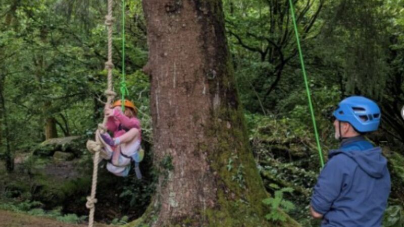 A young girl swinging from a rope swing, supported by a rope and a member of staff holding the rope