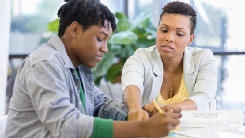 A teenager at home with their mum sat at a table looking at a school book.