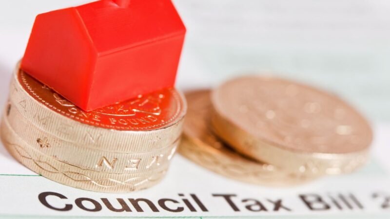 image shows a small house (like a 'Monopoly' house) on the top of a pile of pound coins, on top of a council tax bill.