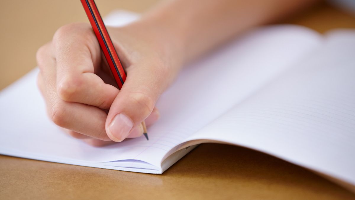 A young pupil writing in their book. Only their hands and a pencil and exercise book in the picture