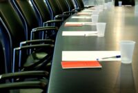 A large meeting desk ready for a Board meeting. Papers are piled neatly in people's places. Seat around the table are unoccupied
