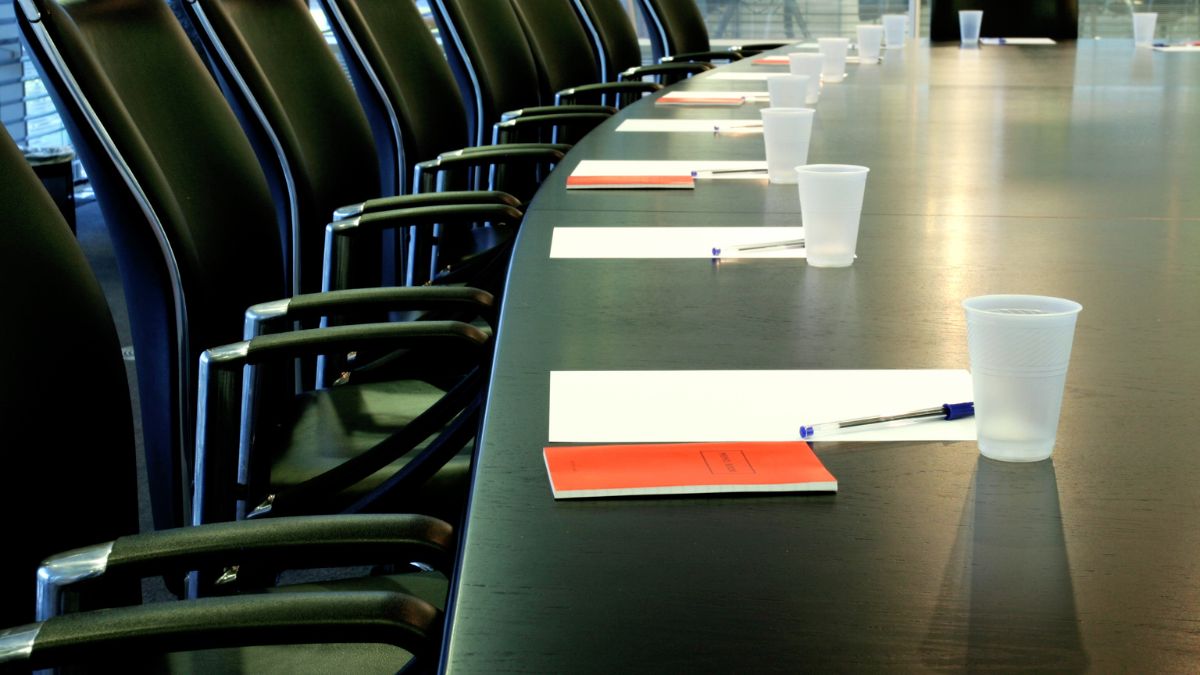 A large meeting desk ready for a Board meeting. Papers are piled neatly in people's places. Seat around the table are unoccupied