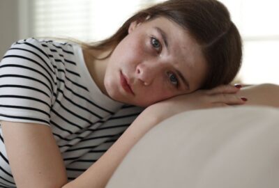 A teenage girl sat with her head resting on her arm, sat on a settee at home, looking anxious
