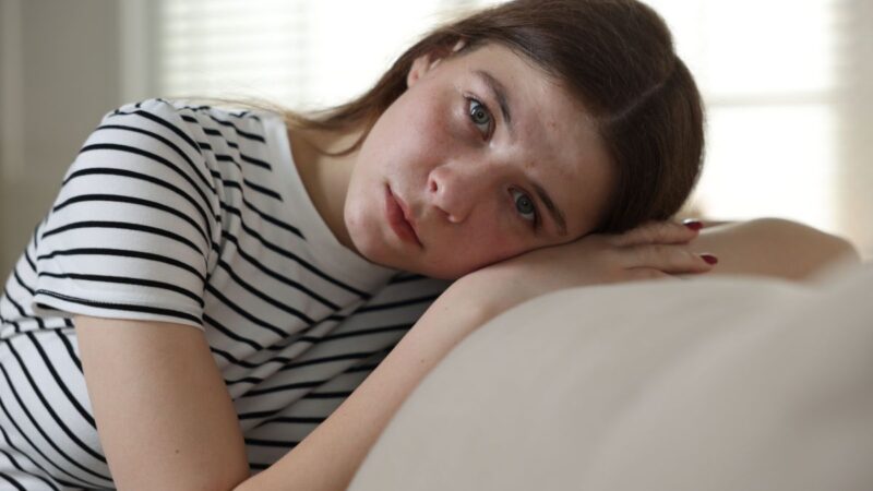 A teenage girl sat with her head resting on her arm, sat on a settee at home, looking anxious