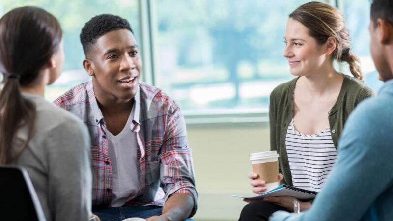 A group of young adults, holding cups of hot drink, chatting, smiling.
