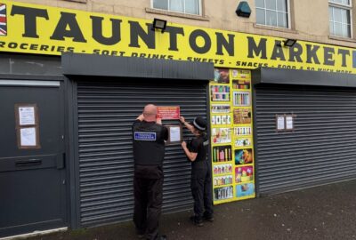 External photo of the shop Taunton Market, with Police and Trading Standards officers putting up a 'closure' notice on the shop's shutters