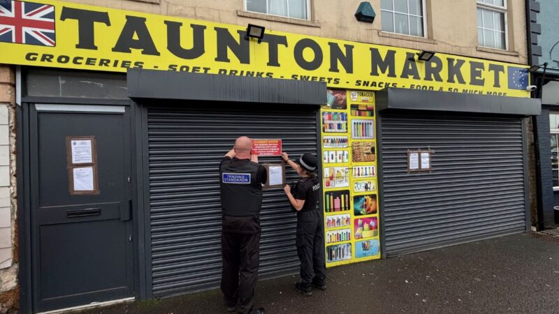 External photo of the shop Taunton Market, with Police and Trading Standards officers putting up a 'closure' notice on the shop's shutters