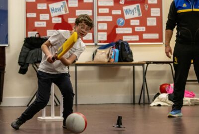 Boy in the classroom playing, (supervised) with a cricket bat and a football, having fun.
